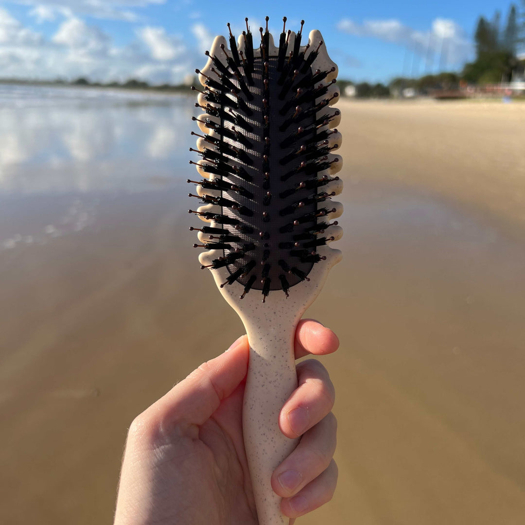 Hand holding a beige curl defining hair brush at the beach