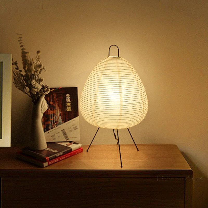 Minimalist Japanese rice paper table lamp glowing softly on a wooden nightstand beside a vase of dried flowers.