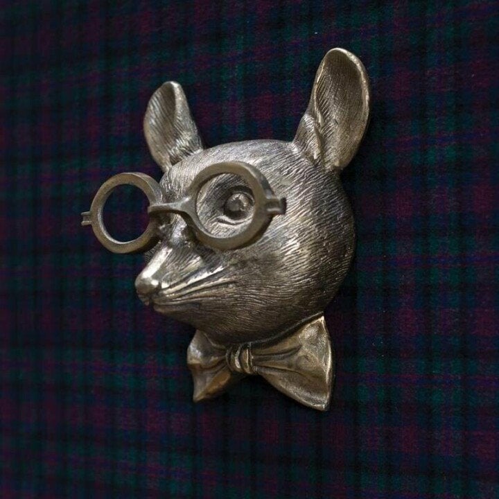 Detailed view of a silver mouse wall sculpture wearing round glasses and a bowtie on a plaid background