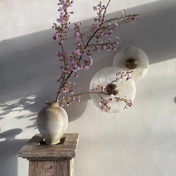 Two round alabaster wall lamps mounted on a neutral wall next to a floral vase.
