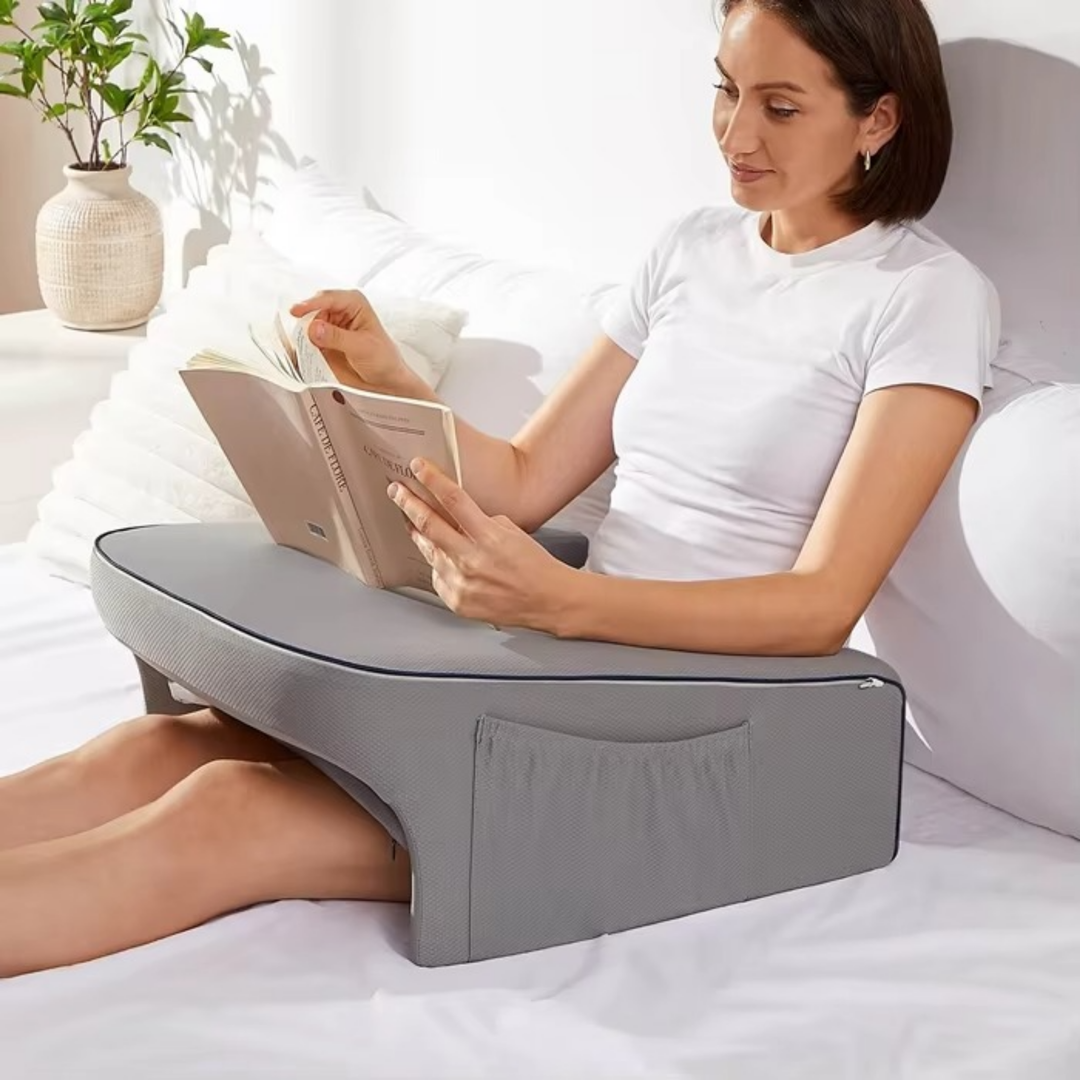Woman reading a book in bed using a grey ergonomic lap pillow with armrests