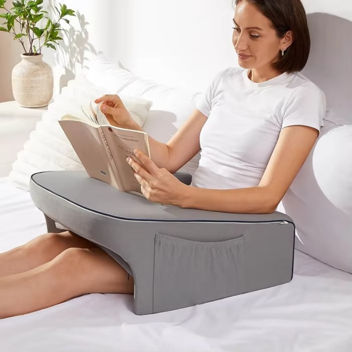 Woman reading a book in bed using a grey ergonomic lap pillow with armrests