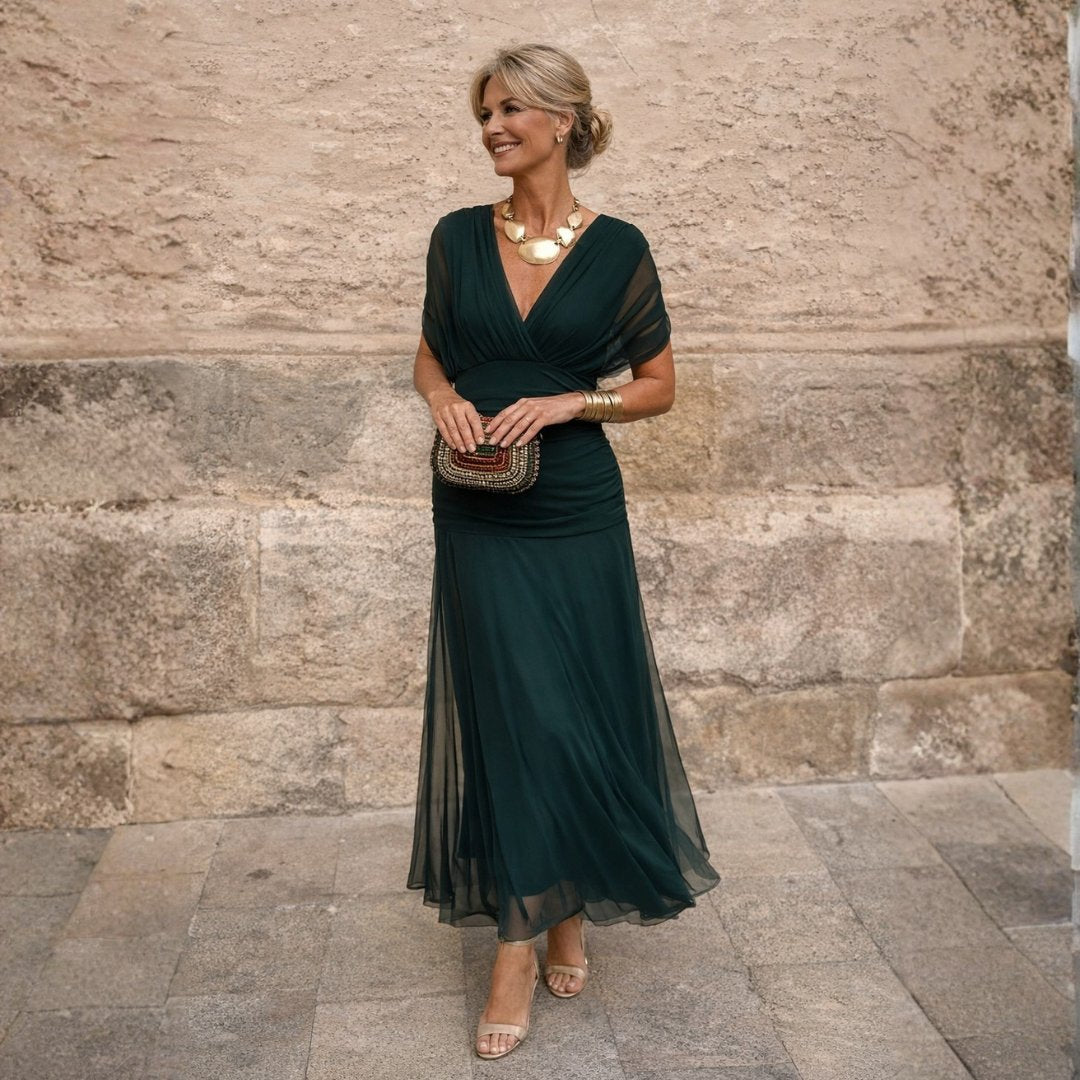 Woman wearing a dark green off-shoulder maxi dress outdoors, accessorised with a statement gold necklace and a clutch.