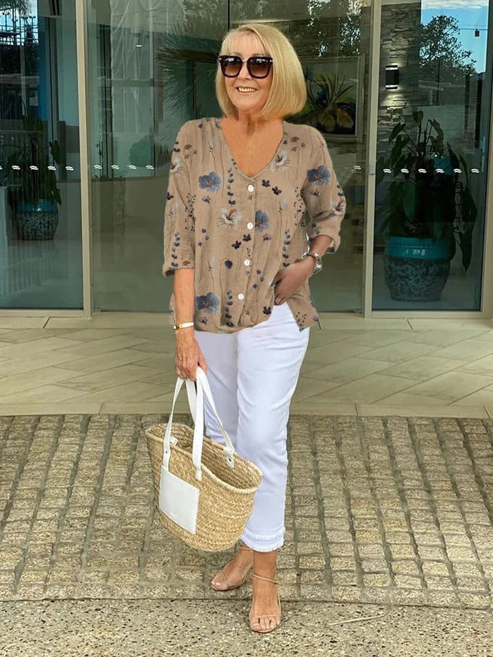 Woman wearing a khaki floral print V-neck blouse outdoors with white trousers and a woven tote bag