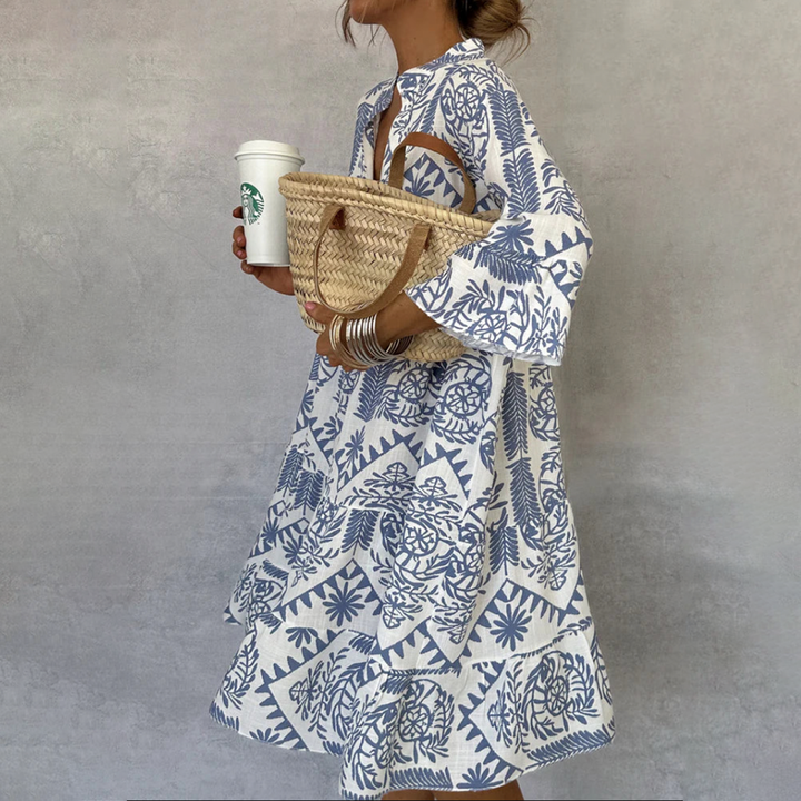 Woman wearing a blue and white patterned bohemian midi dress with long sleeves holding a straw bag and coffee cup