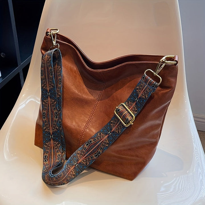 Brown oversized retro shoulder bag with patterned strap resting on a white chair