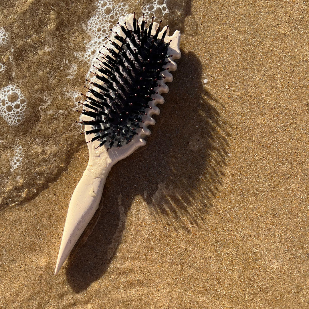 Beige curl defining hair brush resting on sandy beach near water