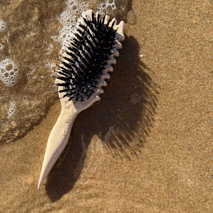 Beige curl defining hair brush resting on sandy beach near water