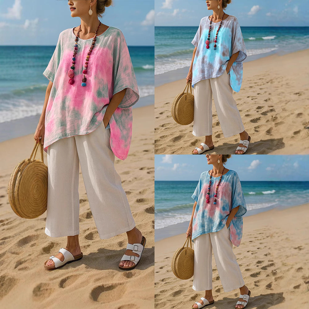 A woman wearing a colorful tunic top and white trousers on a sandy beach.