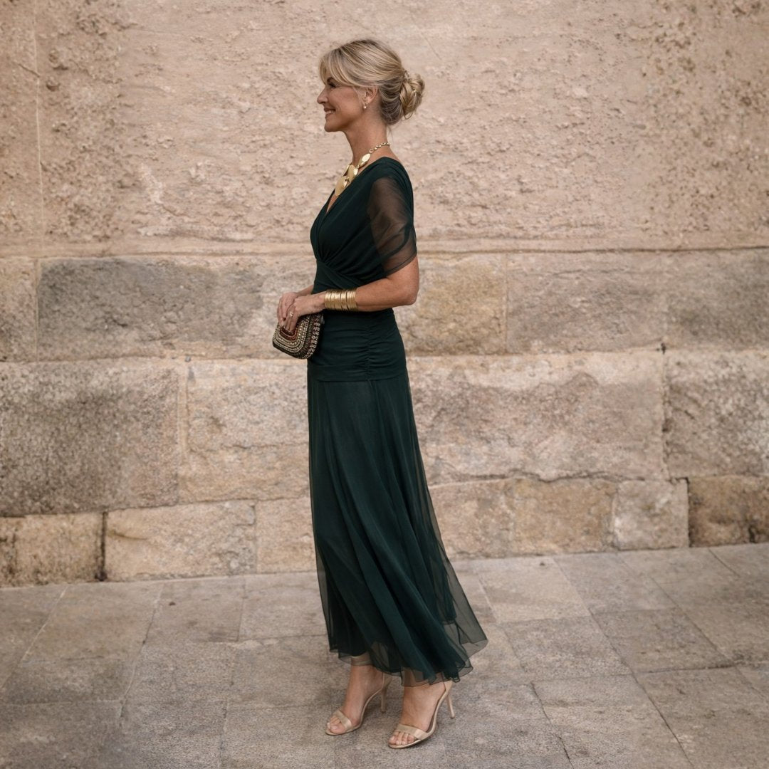 Side profile of a woman showcasing the draped off-shoulder bodice and flowing maxi skirt of the dark green dress.