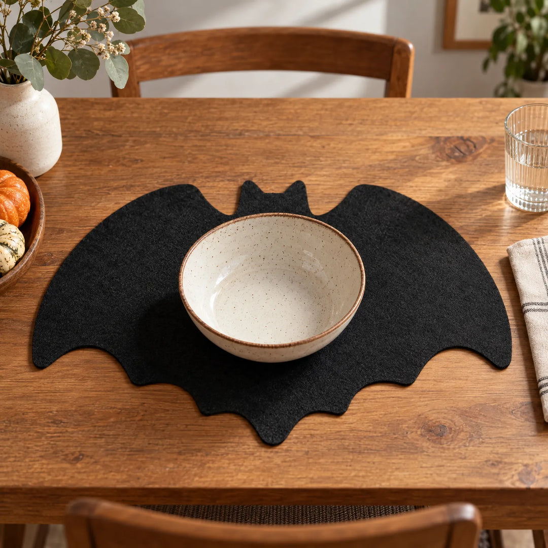 Black bat-shaped felt placemat holding a ceramic bowl on a wooden dining table