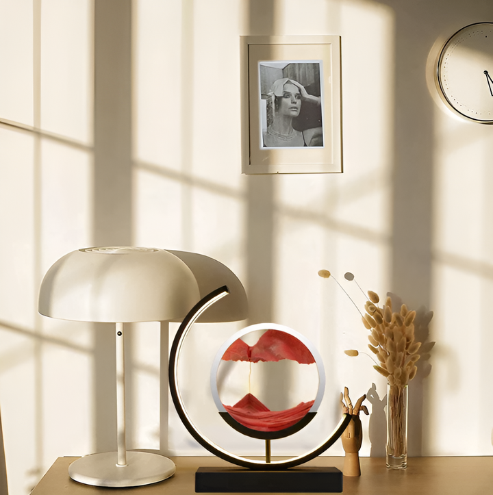 Illuminated sand art lamp placed on a wooden desk next to a white table lamp and vase.