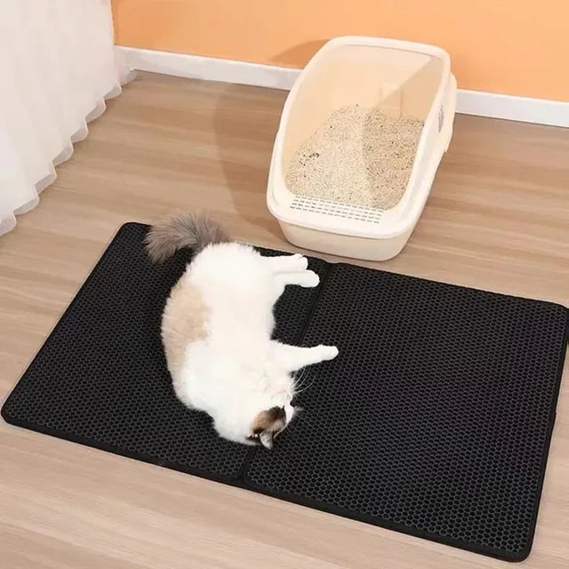 Black double-layer cat litter mat placed on wooden floor next to litter box with resting cat