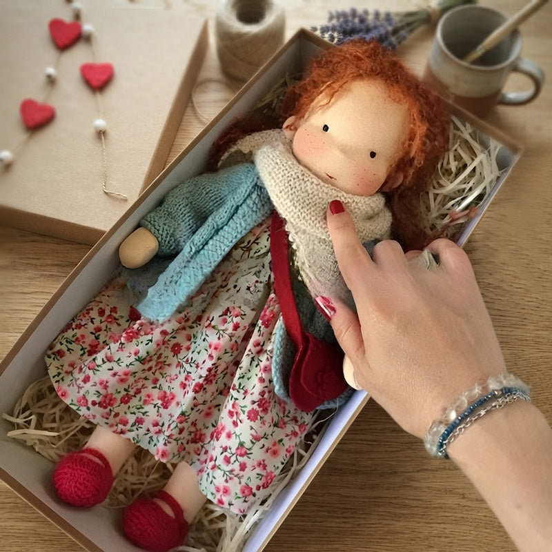 Handmade Waldorf doll nestled inside a gift box with a hand pointing to it