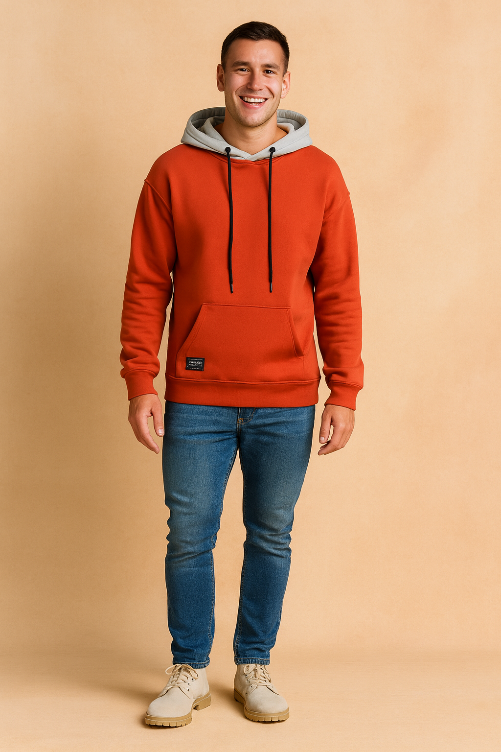 Smiling man wearing an orange oversize hoodie paired with blue jeans and casual boots against a neutral background.