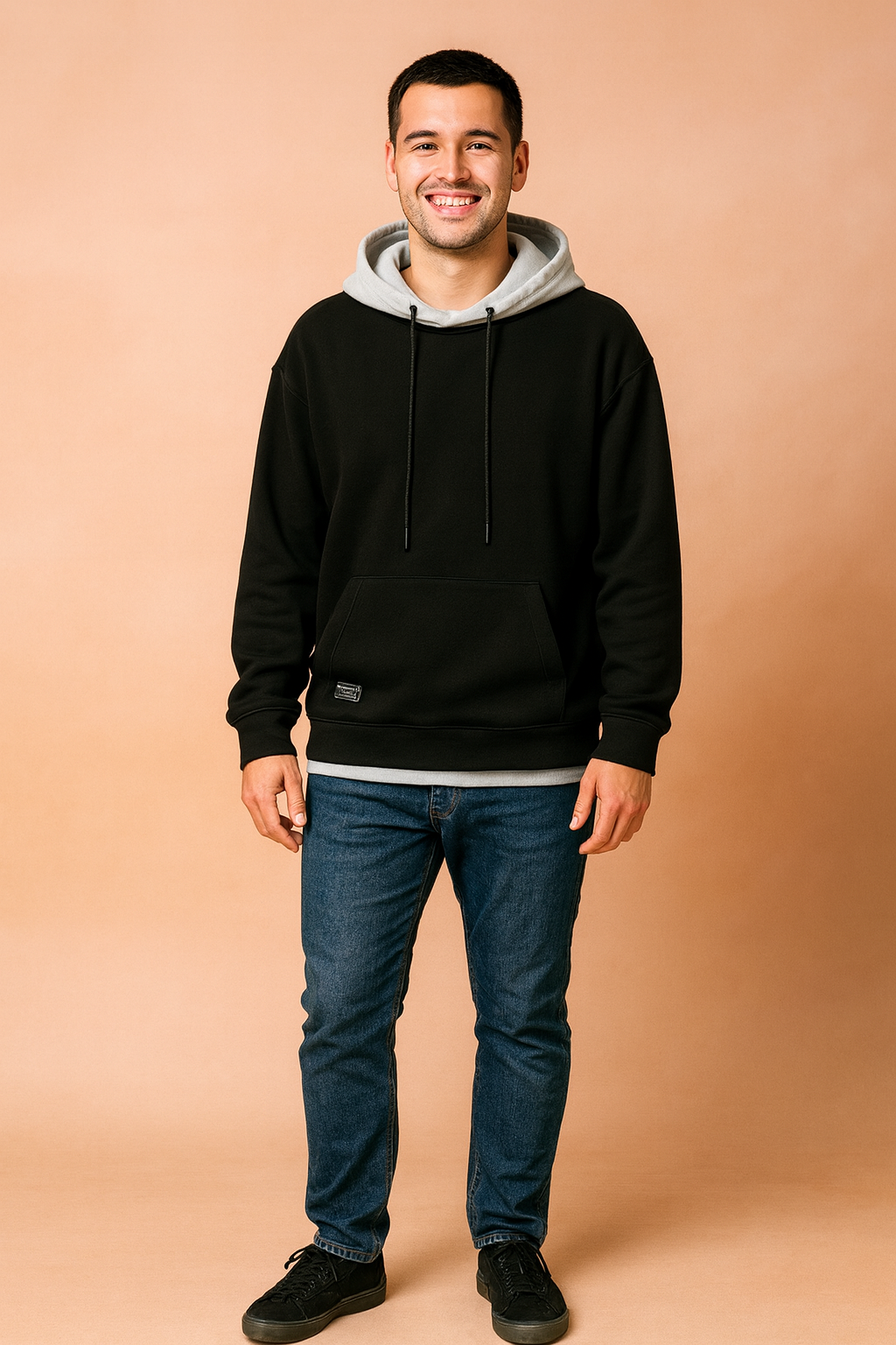 Smiling man wearing a black oversize hoodie paired with blue jeans and black sneakers against a neutral background.