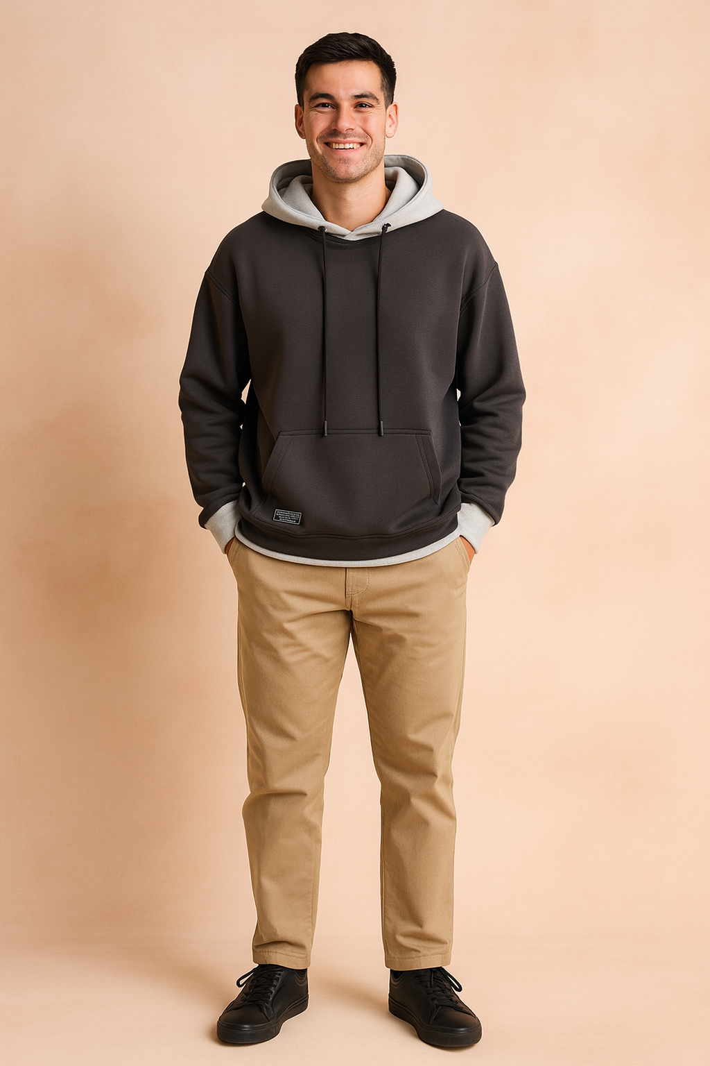 Smiling man wearing a dark gray oversize hoodie with hands in pockets, paired with khaki trousers and black sneakers.