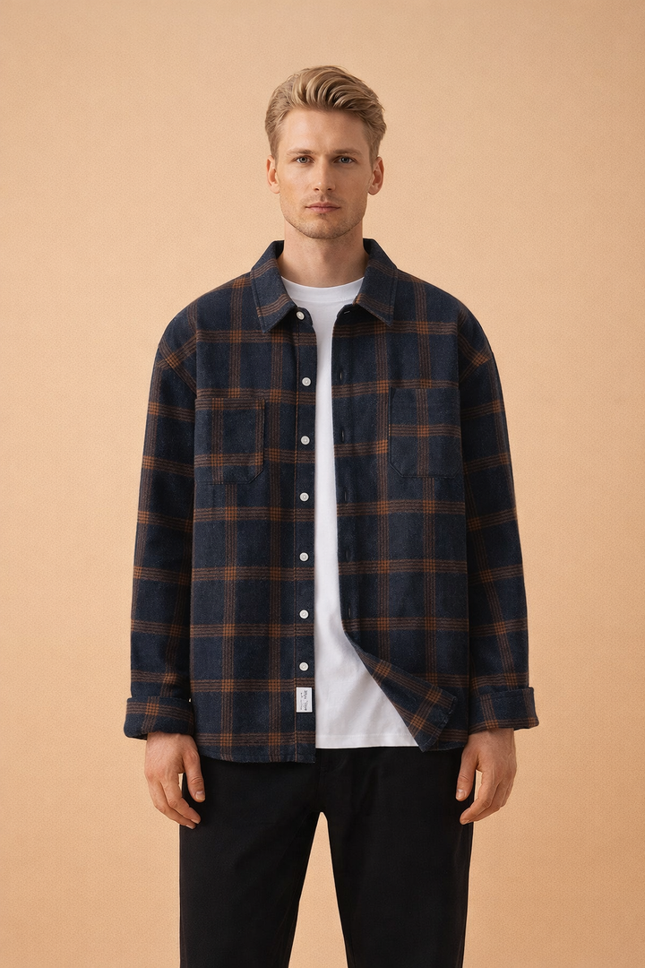 Blonde male model wearing a navy blue and brown checked long-sleeve overshirt over a white t-shirt and black trousers.