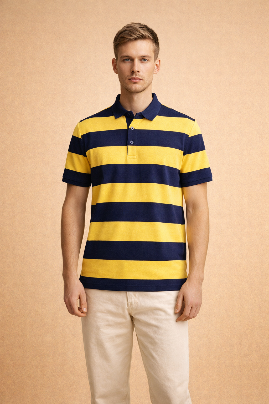 Blonde male model wearing a yellow and navy striped short-sleeve polo shirt and neutral trousers against a warm backdrop.