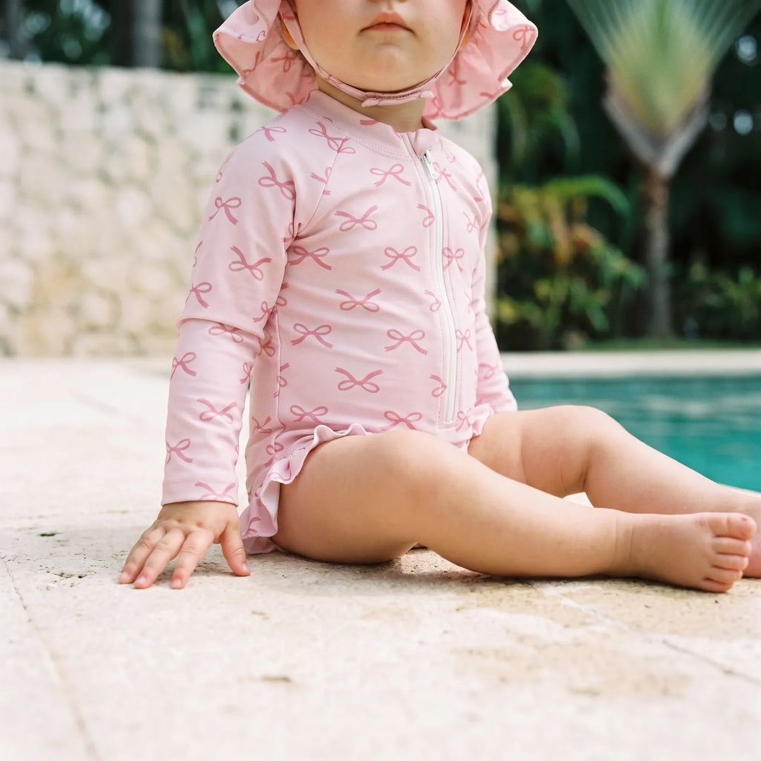 Toddler sitting by the pool wearing a pink bow-print long-sleeve swimsuit and matching sun hat
