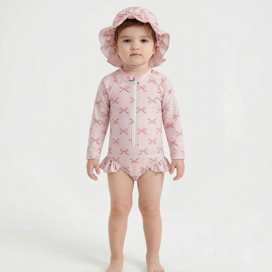 Full-length front view of a baby wearing a pink bow-print long-sleeve swimsuit and sun hat