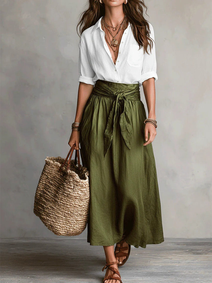 Woman wearing a white button-down shirt and green tie-waist midi skirt holding a straw tote bag