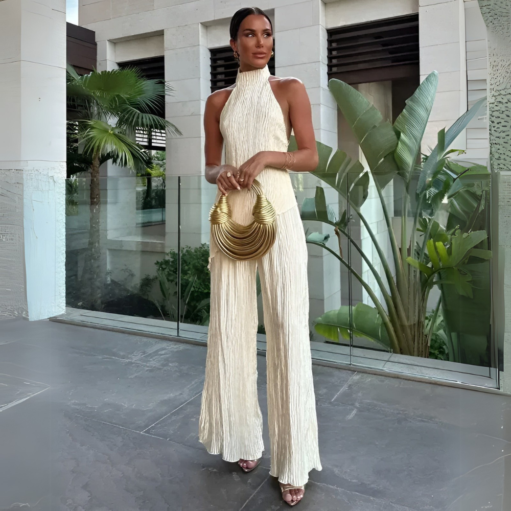 Model standing outdoors wearing a beige pleated halter wide leg jumpsuit holding a gold handbag