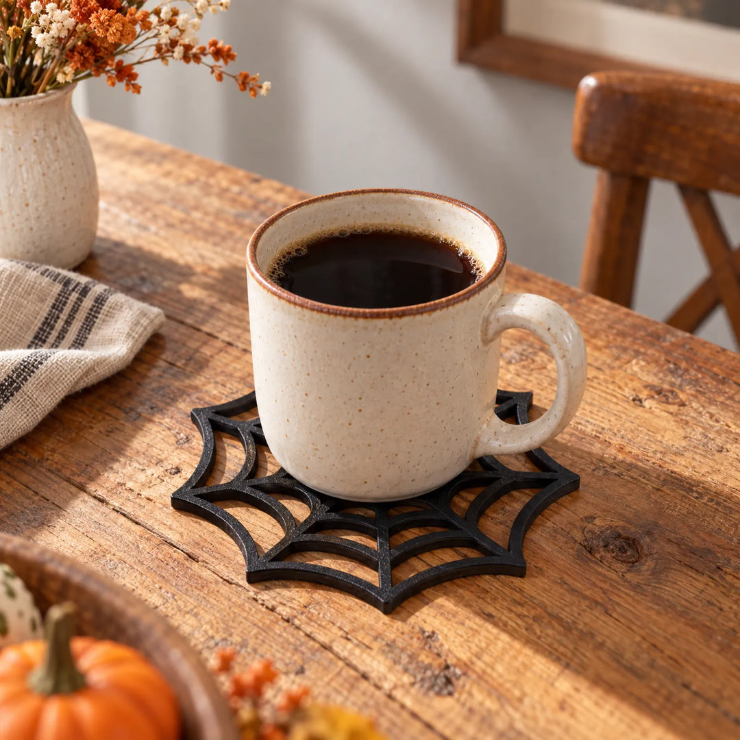 Black spiderweb drink coaster holding a ceramic coffee mug on a rustic wooden table
