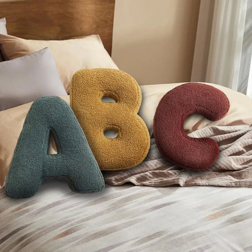 Textured letter cushions shaped like A, B, and C arranged on a bed with neutral pillows.