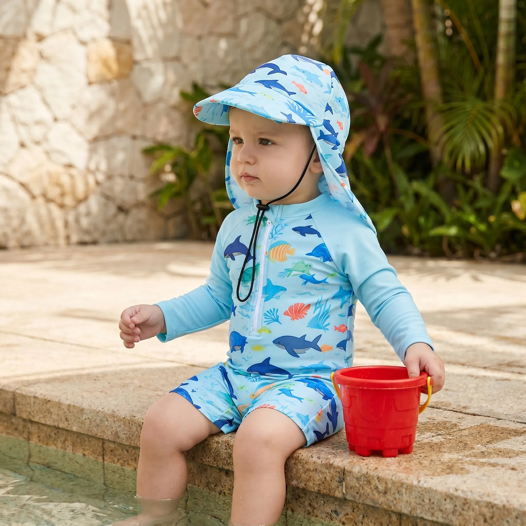 Toddler sitting by the pool wearing light blue marine print swimming romper and sun hat