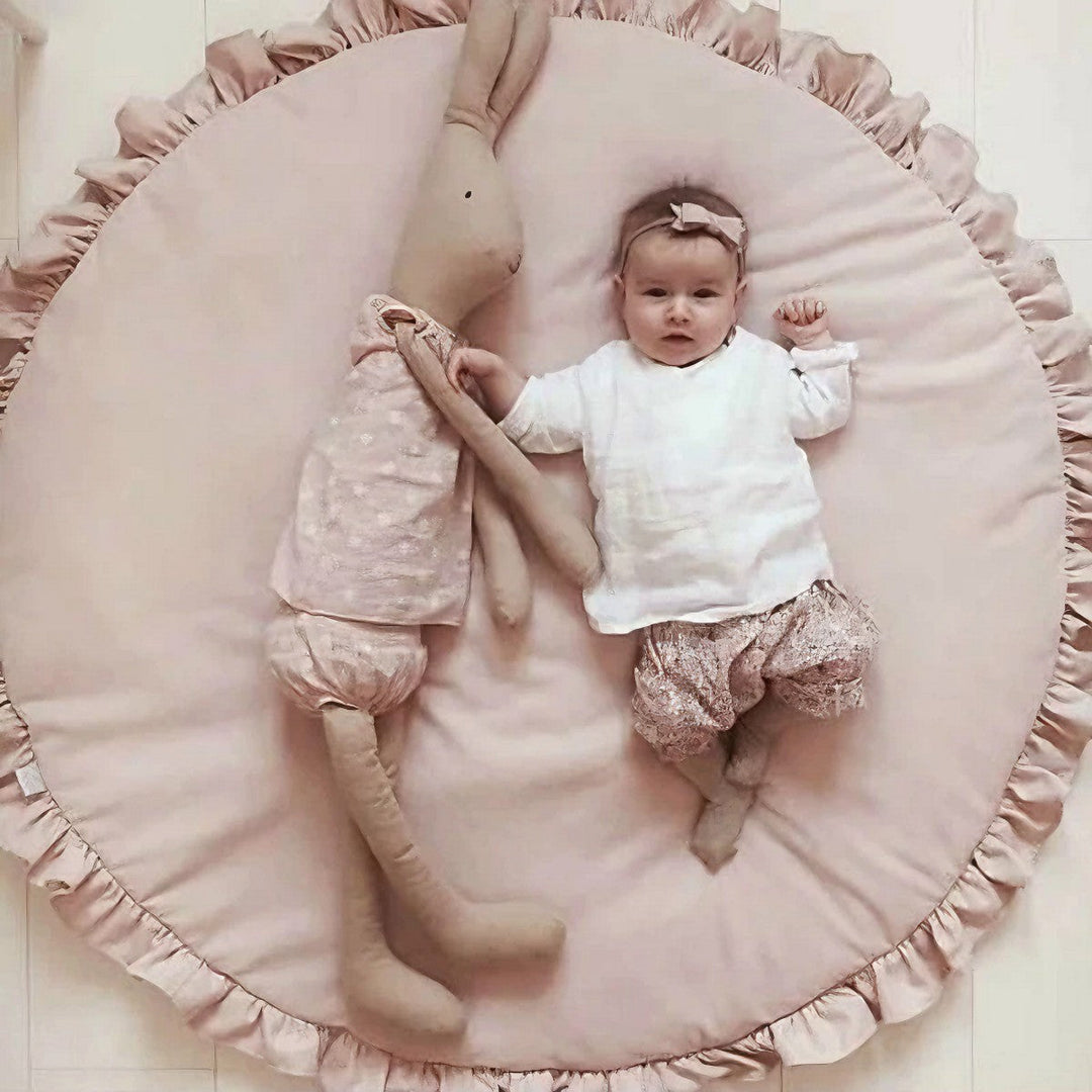 Baby lying comfortably on a soft pink round play mat with decorative ruffles alongside a large stuffed bunny toy.