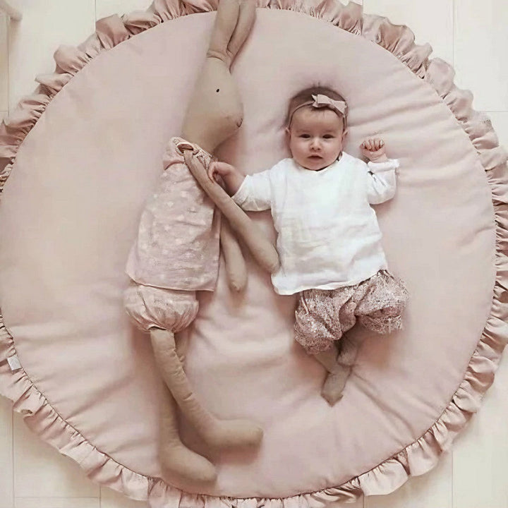 Baby lying comfortably on a soft pink round play mat with decorative ruffles alongside a large stuffed bunny toy.