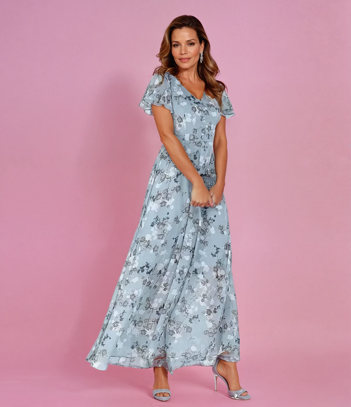 Model wearing blue floral ruffle sleeve maxi dress with silver heels against a pink background