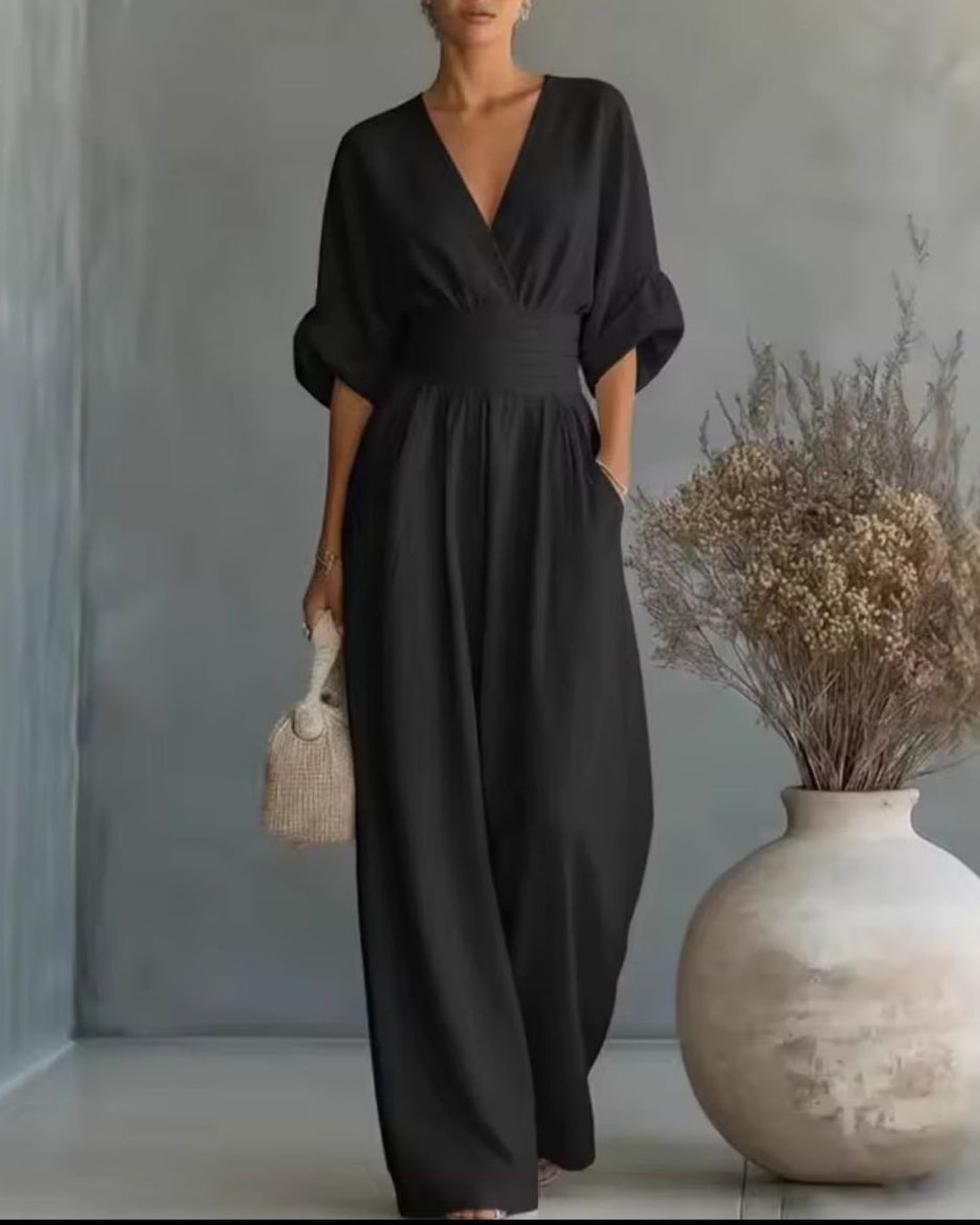 Woman wearing the black V-neck long sleeve jumpsuit holding a woven bag