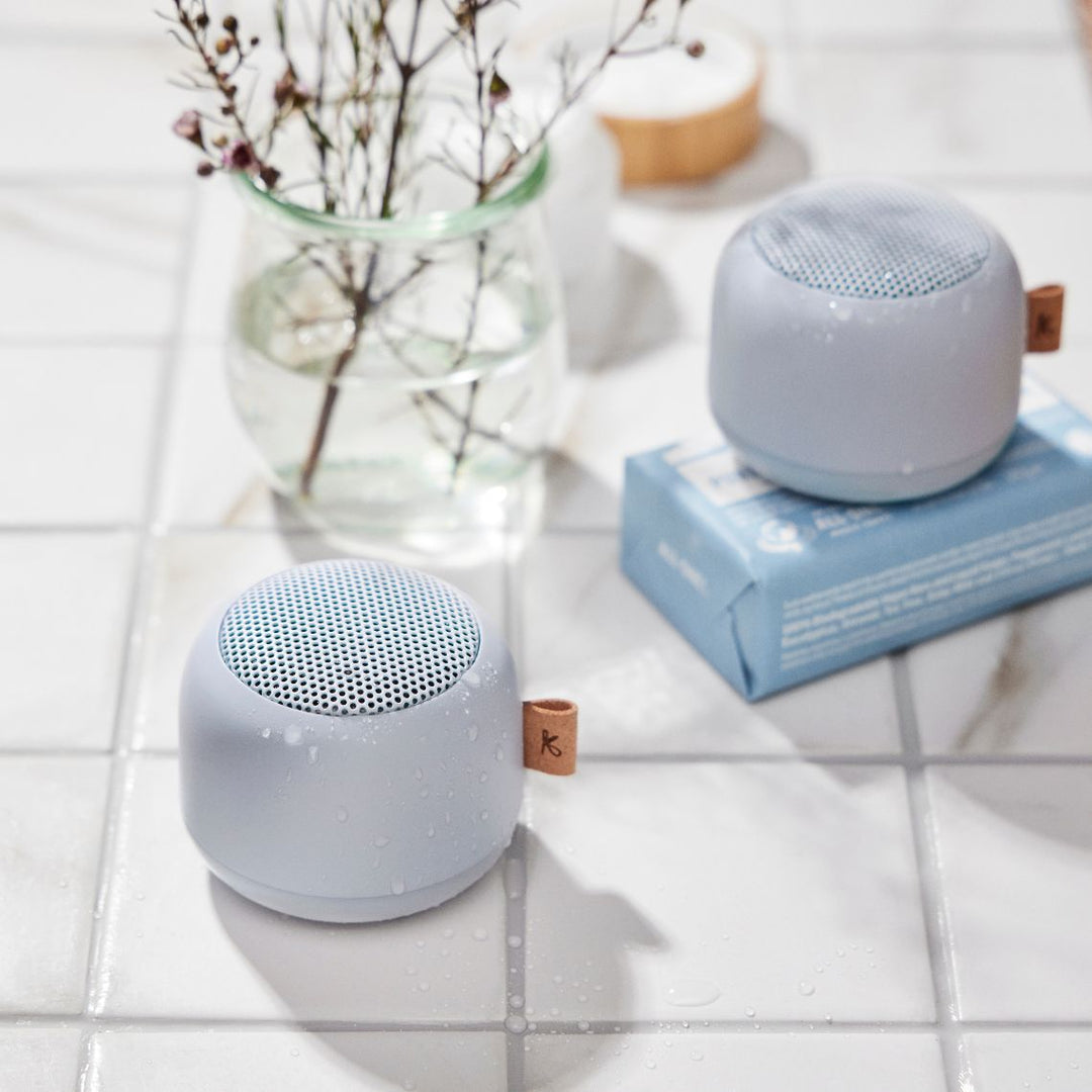 Cloudy blue waterproof bluetooth speaker next to a glass vase with plants on a tiled surface