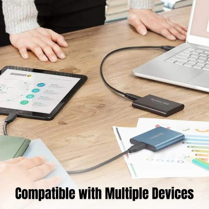 Portable external SSD hard drives connected to a laptop and tablet on a wooden desk.