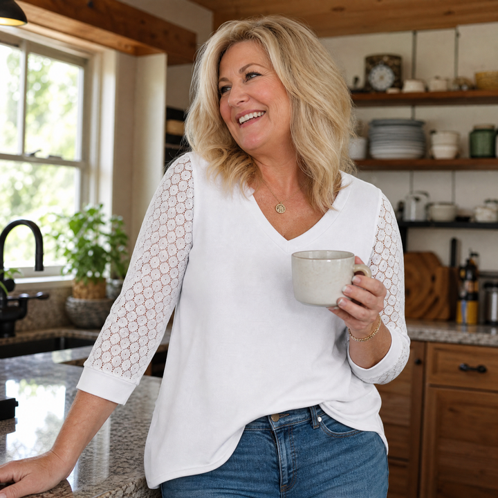 Smiling woman holding a mug in a kitchen while wearing a white V-neck top with lace sleeves.