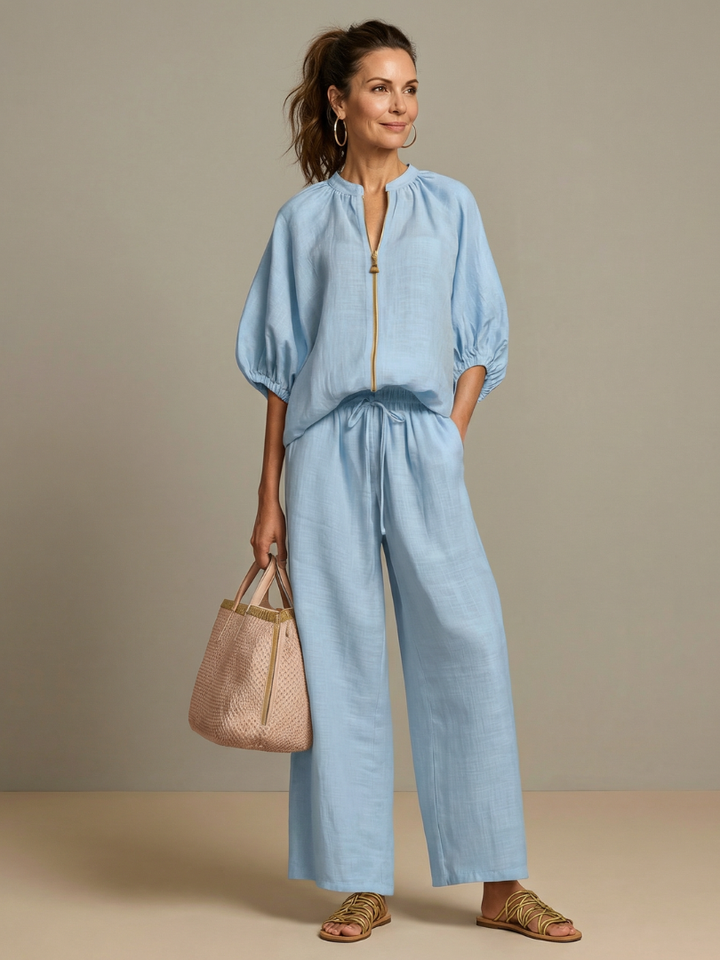 Woman wearing light blue co-ord set with wide leg pants and zip top holding a woven bag