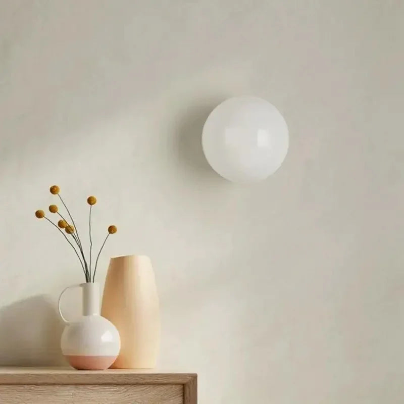 Minimalist frosted glass wall lamp mounted on a neutral wall next to a vase with dried flowers