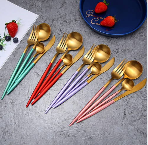 Assorted gold-plated cutlery sets with colorful handles displayed on a dark surface alongside a plate and strawberries