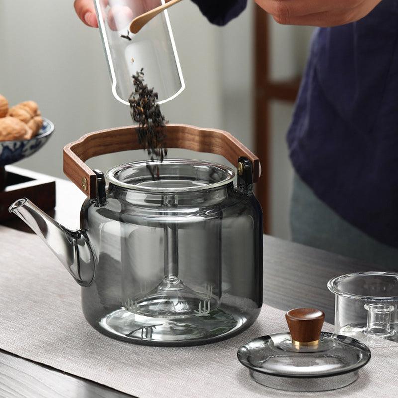 Person pouring tea leaves into the smoked glass teapot with wooden accents