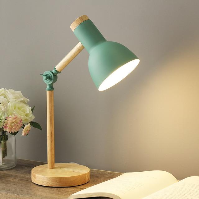 Green Scandinavian wooden table lamp on a wooden desk next to a book and flowers