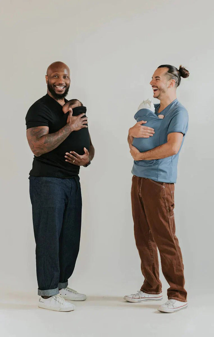 Two fathers smiling while wearing infants in baby carrier shirts