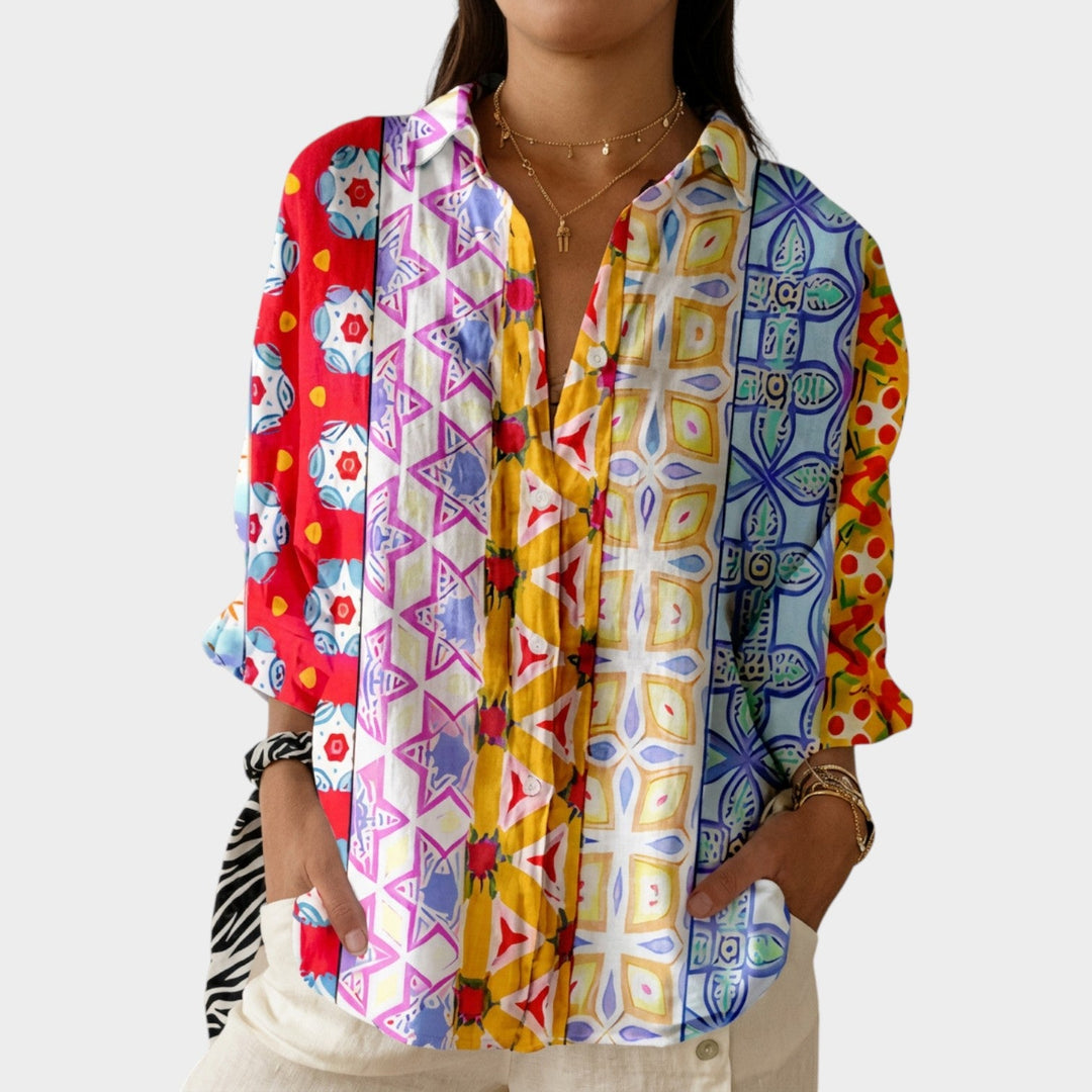 Woman wearing a vibrant multi-colored patterned button-up blouse with layered gold necklaces.