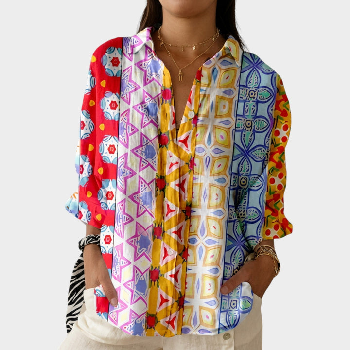 Woman wearing a vibrant multi-colored patterned button-up blouse with layered gold necklaces.
