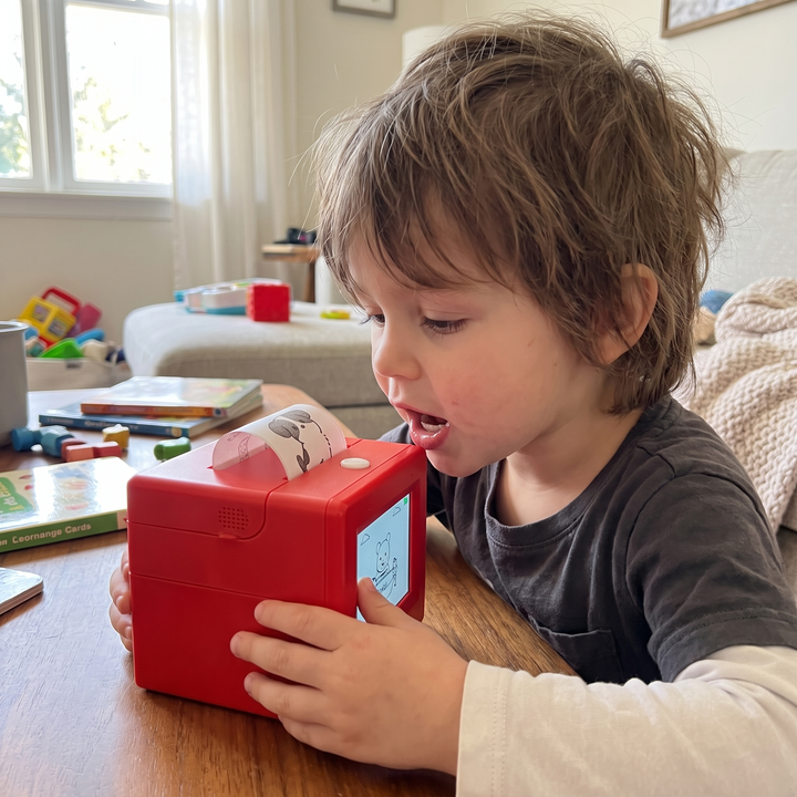 Young child talking to a red AI voice-activated mini sticker printer at a wooden table