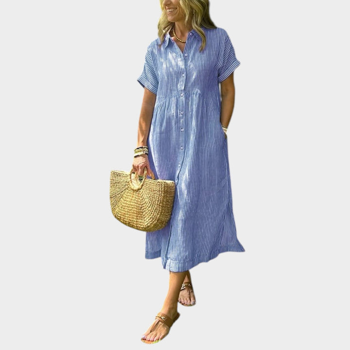 Woman wearing a blue striped maxi shirt dress holding a straw bag and sandals
