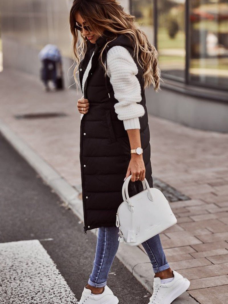 Side view of woman posing in a black knee-length quilted bodywarmer holding a white handbag