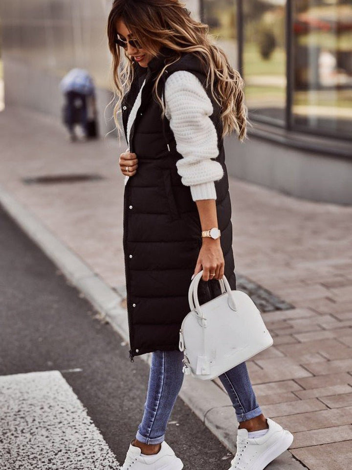 Side view of woman posing in a black knee-length quilted bodywarmer holding a white handbag