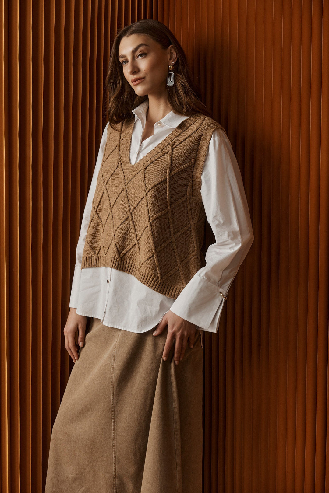 Model wearing a women's mocha V-neck knit vest layered over a white shirt against a textured backdrop